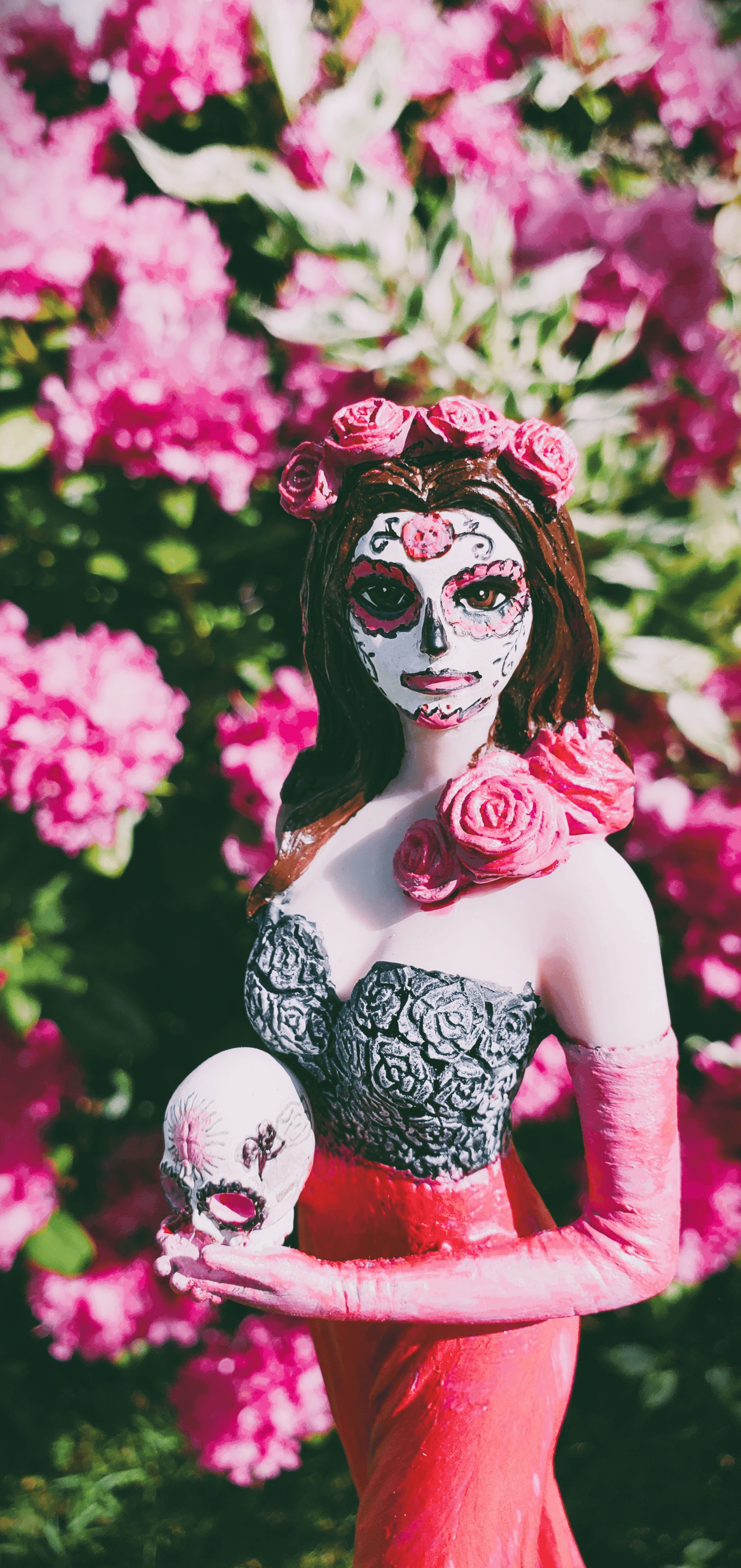 Woman with dark hair wearing catrina makeup and holding a painted skull, surrounded by vibrant pink flowers