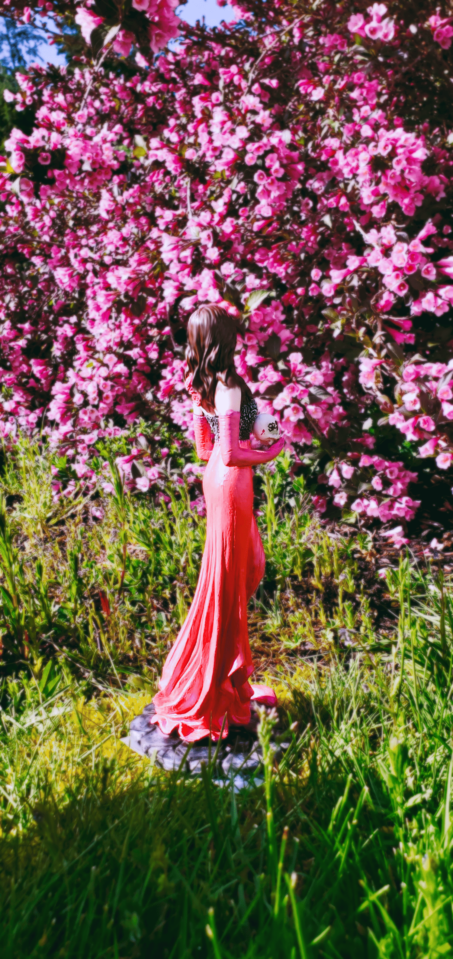 Woman with dark hair wearing catrina makeup and holding a painted skull, surrounded by vibrant pink flowers